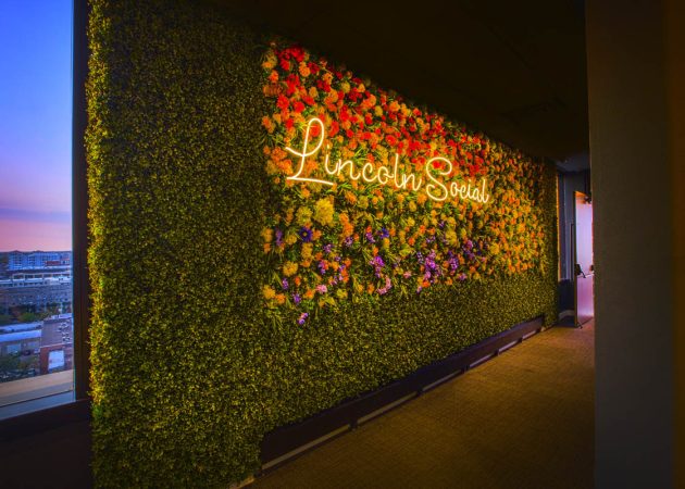 A vibrant floral wall with a neon sign that reads 'Lincoln Social' illuminated in warm light.