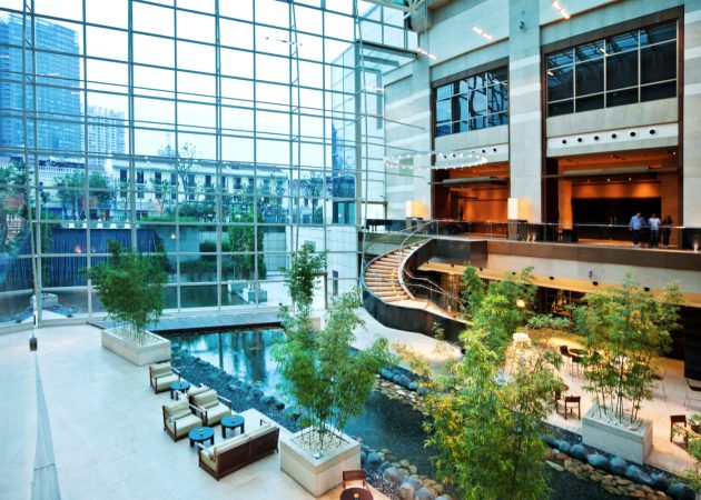 Interior view of a modern lobby featuring glass walls, seating areas, and a water feature with plants.
