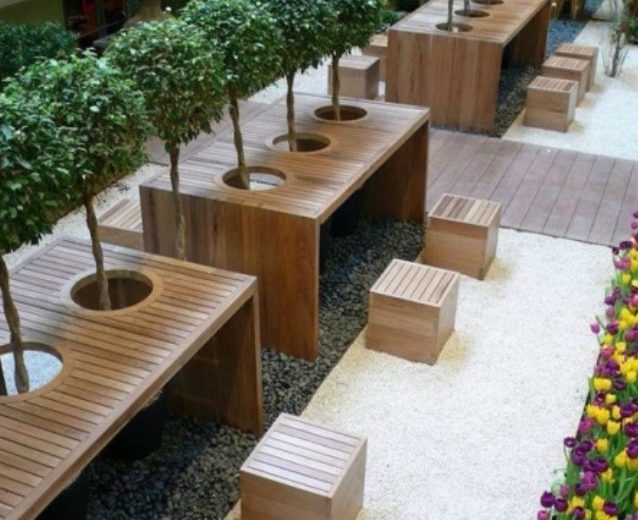 Stylish outdoor seating area featuring wooden tables, planters, and decorative trees in a landscaped environment.