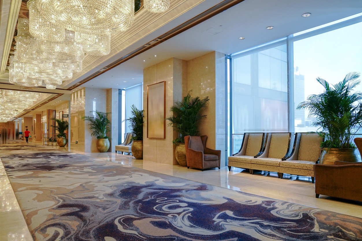 Interior view of a hotel corridor featuring chandeliers, seating, and decorative plants.