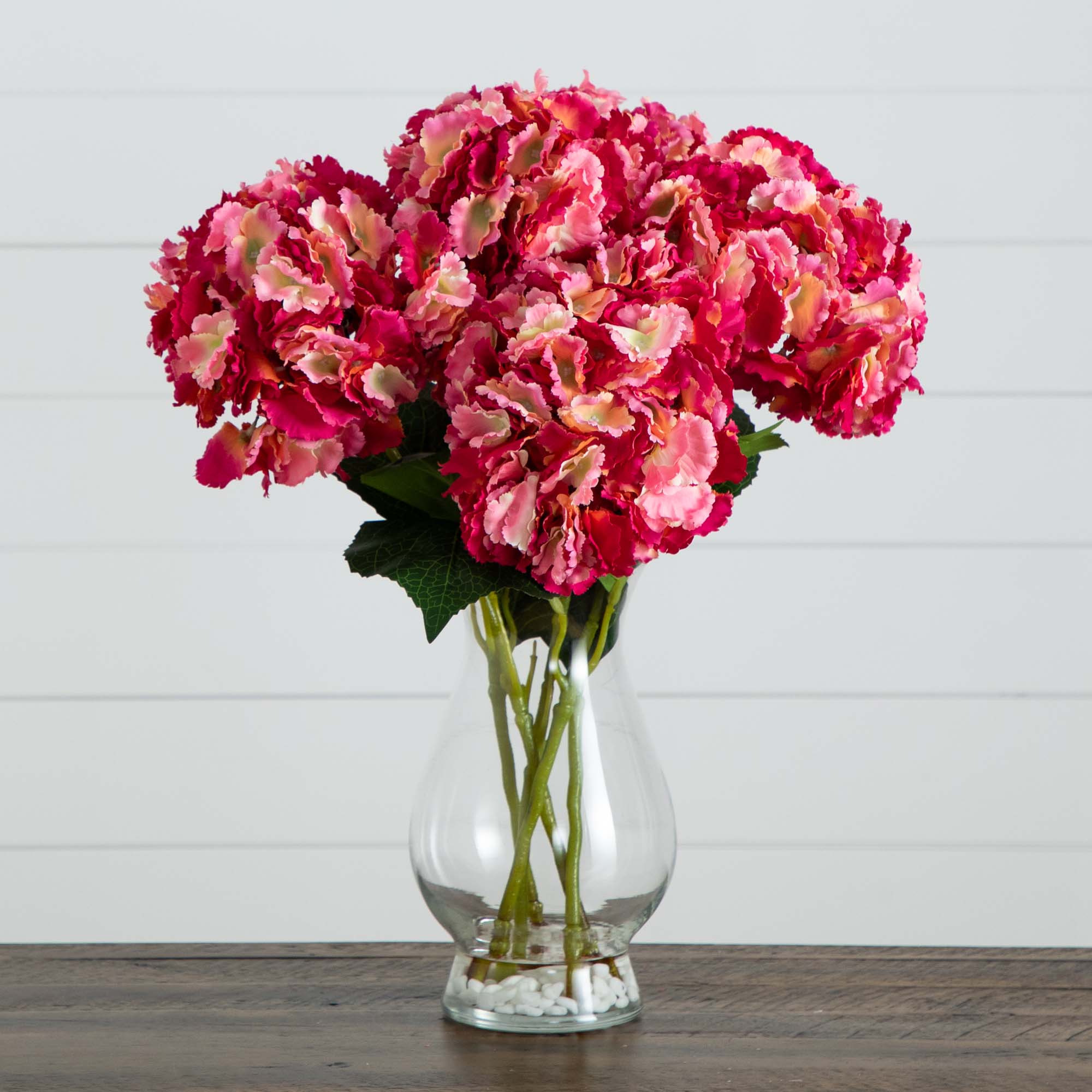 Vibrant artificial pink hydrangea stems fill a clear glass vase on a wooden surface against a white shiplap wall.