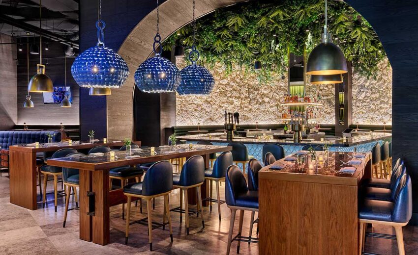 Arched bar interior with leafy green wall texture, blue pendant lights overhead, and polished countertops with glassware under warm lighting.