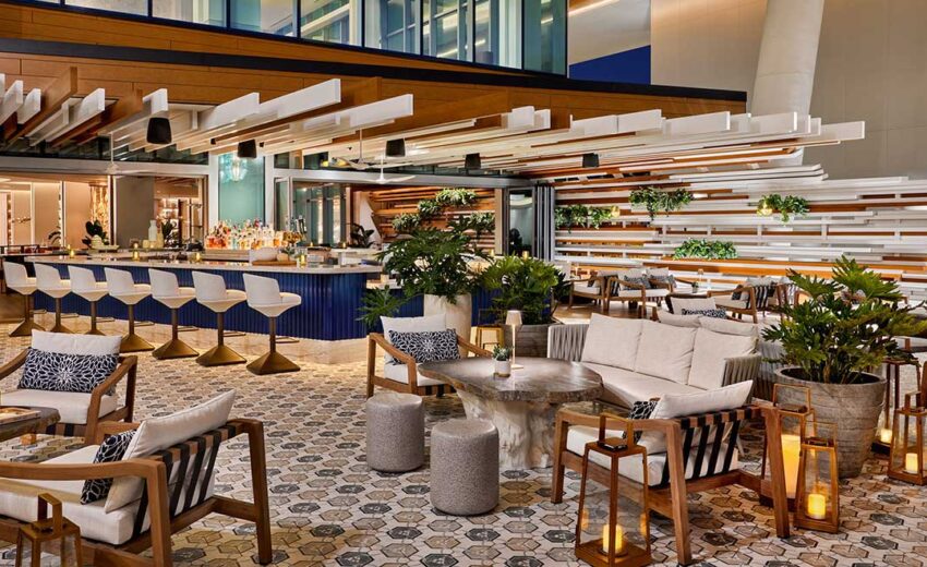 Warmly lit outdoor bar area with a blue counter, white ceiling slats, patterned flooring, and potted replica plants creating a lush, inviting atmosphere.